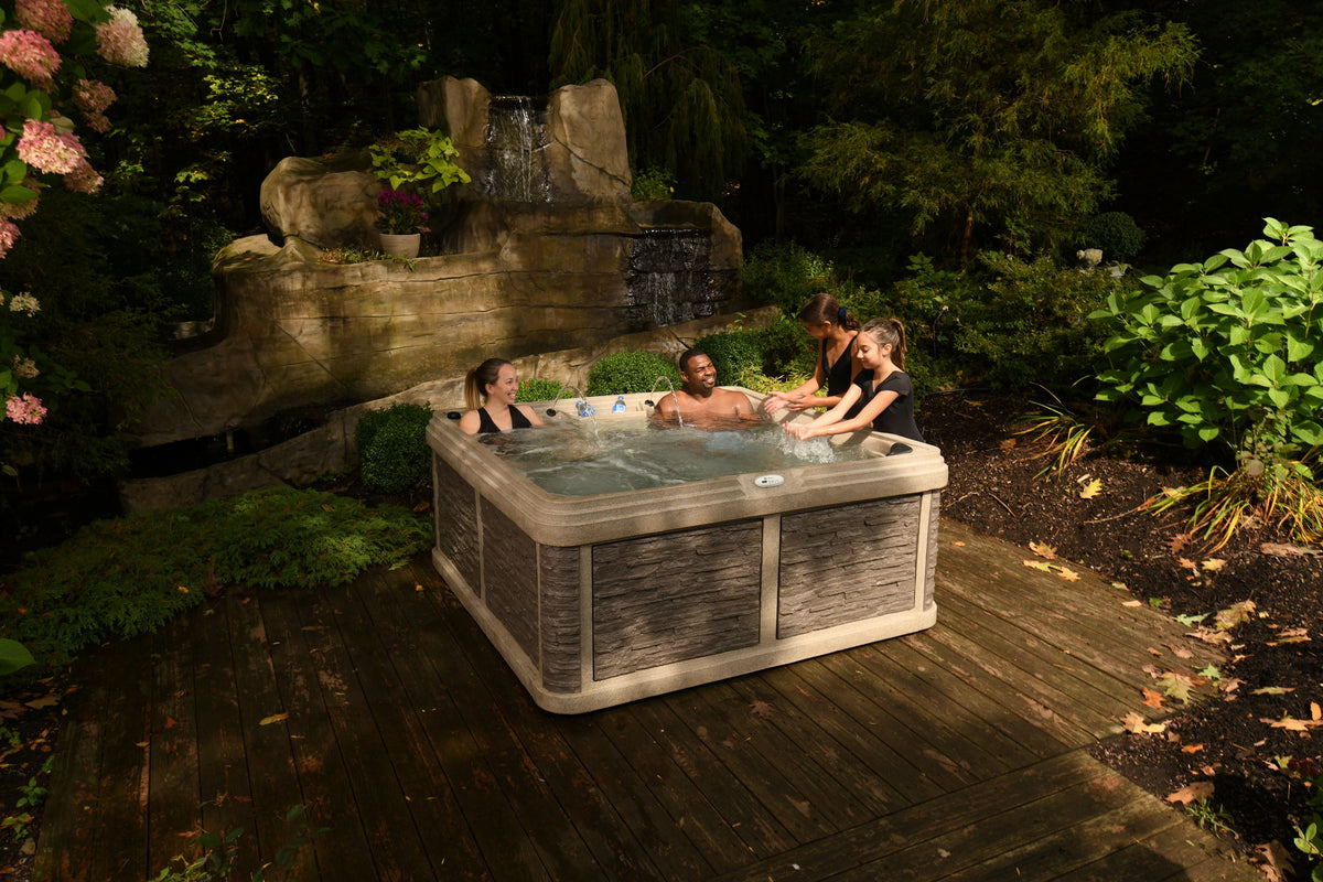 Mesa outdoor hot tub with gray wrap siding sits on a wooden deck in a landscaped garden setting, with three people sitting and relaxing inside. A stone waterfall feature is visible in the background.