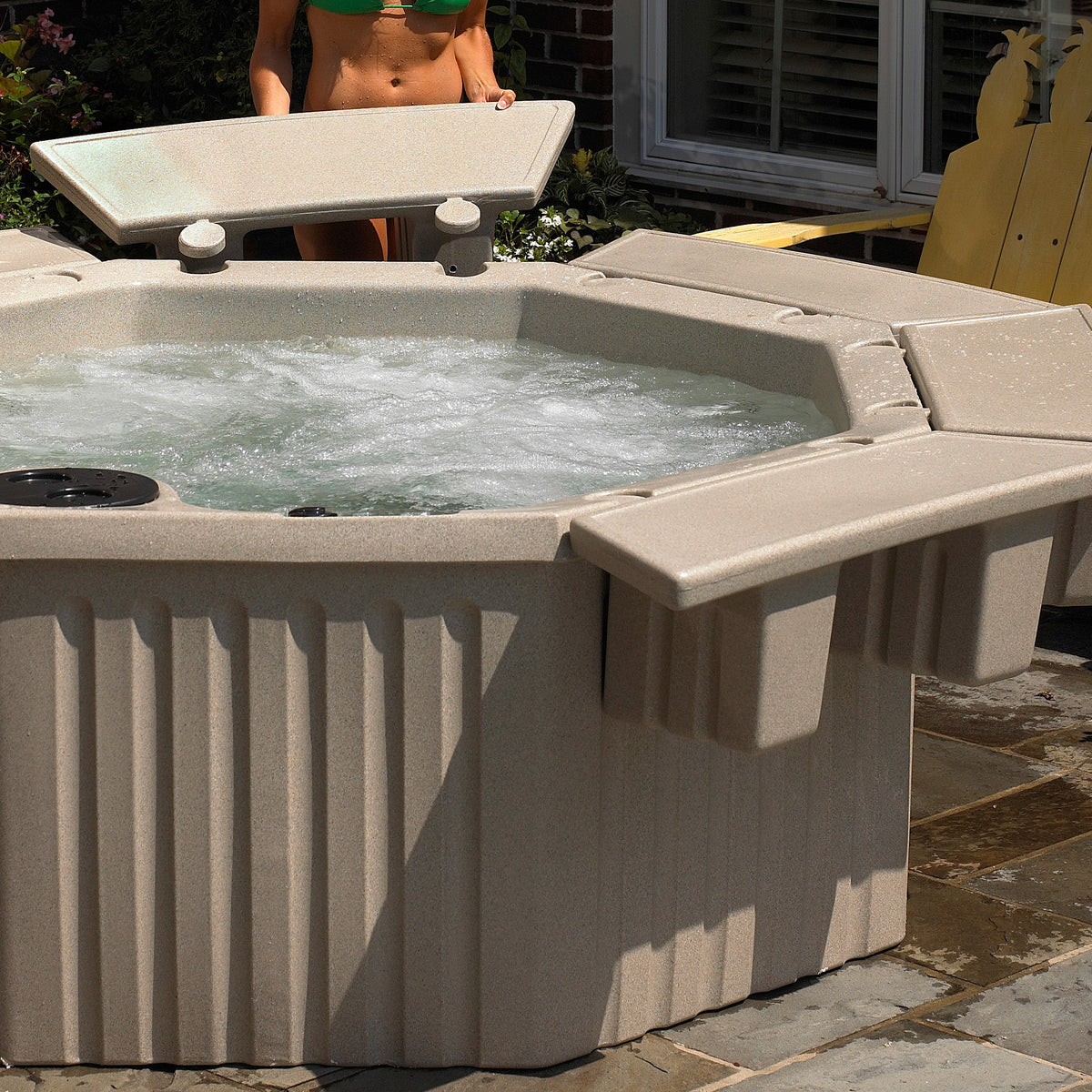 A beige outdoor hot tub with bubbling water, shown on a patio with a person partially visible behind it and a yellow chair in the background.