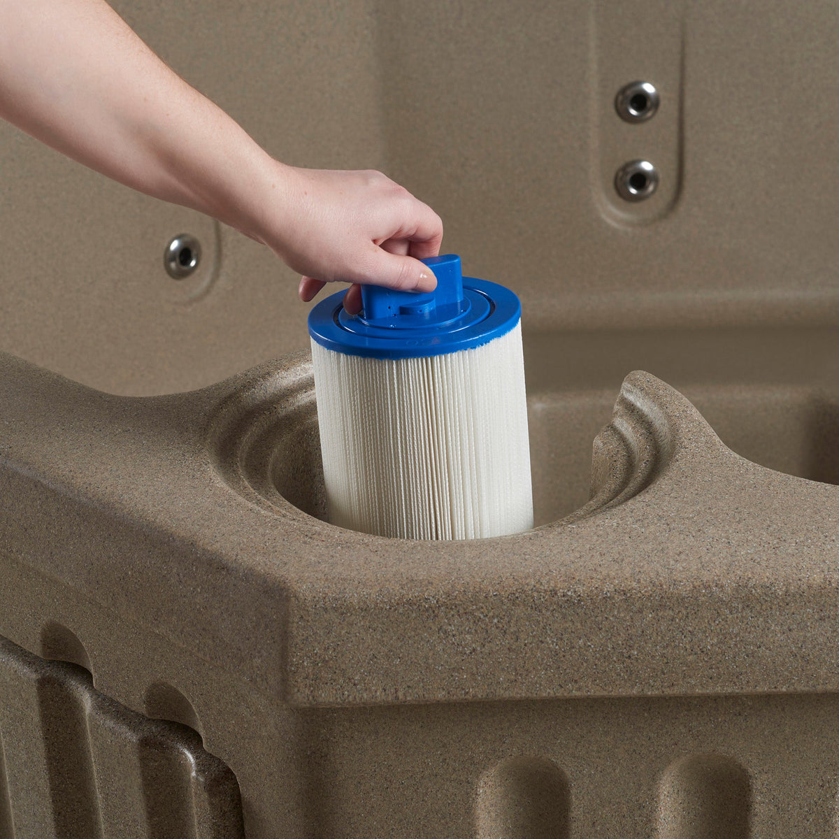 A hand inserts a white pleated filter cartridge with a blue top into a beige spa.