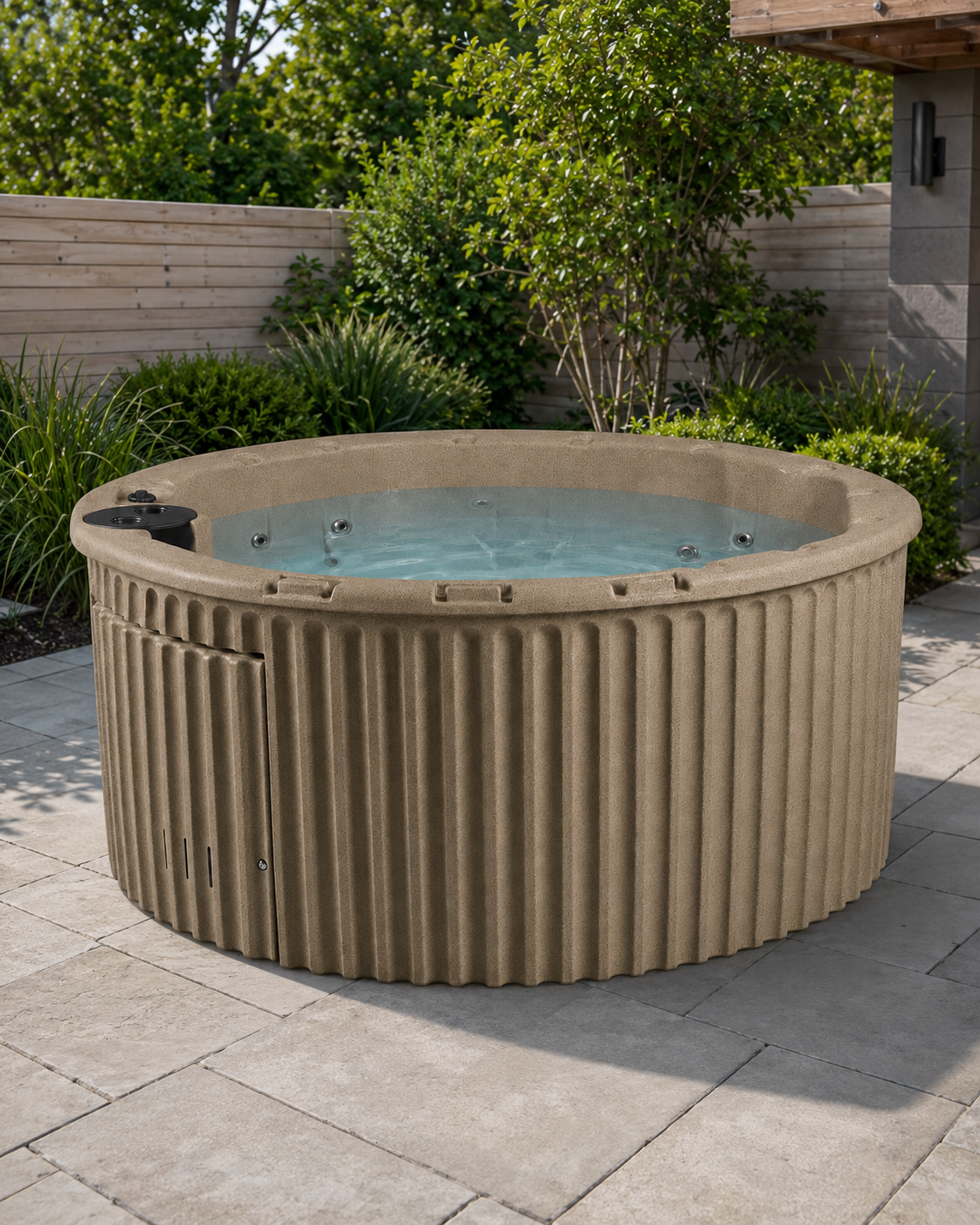 A round outdoor hot tub with a ribbed beige exterior and light blue water, situated on a stone patio with green shrubs and a wooden fence in the background.