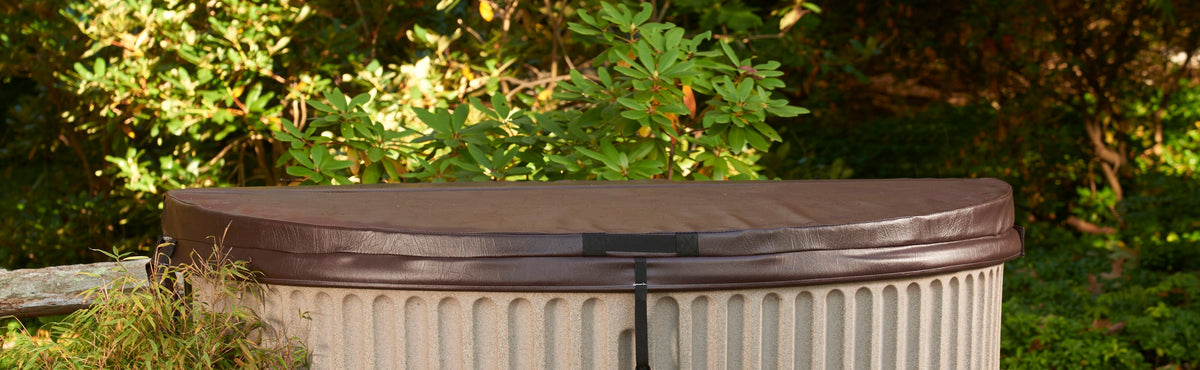 A round outdoor hot tub with a dark brown lid and ribbed beige body, set against a background of green garden foliage.