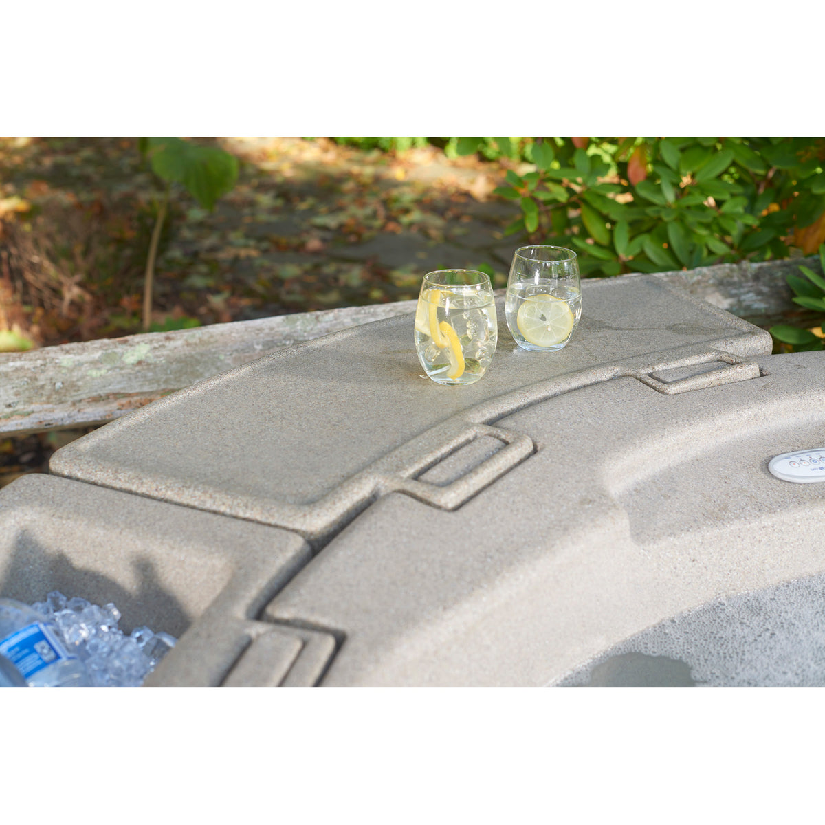 A gray molded outdoor hot tub tray accessory with, shown outdoors on a stone surface with two clear drinking glasses placed on top. The background includes blurred greenery and ground.