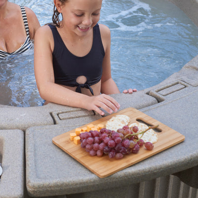 A young girl in a navy swimsuit leans over the edge of a hot tub, smiling as a wooden serving board with grapes and cheese sits on the water edge. Another person is partially visible in the water at left.