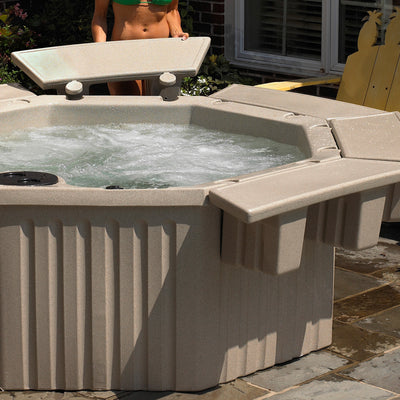 A beige outdoor hot tub with bubbling water, shown on a patio with a person partially visible behind it and a yellow chair in the background.