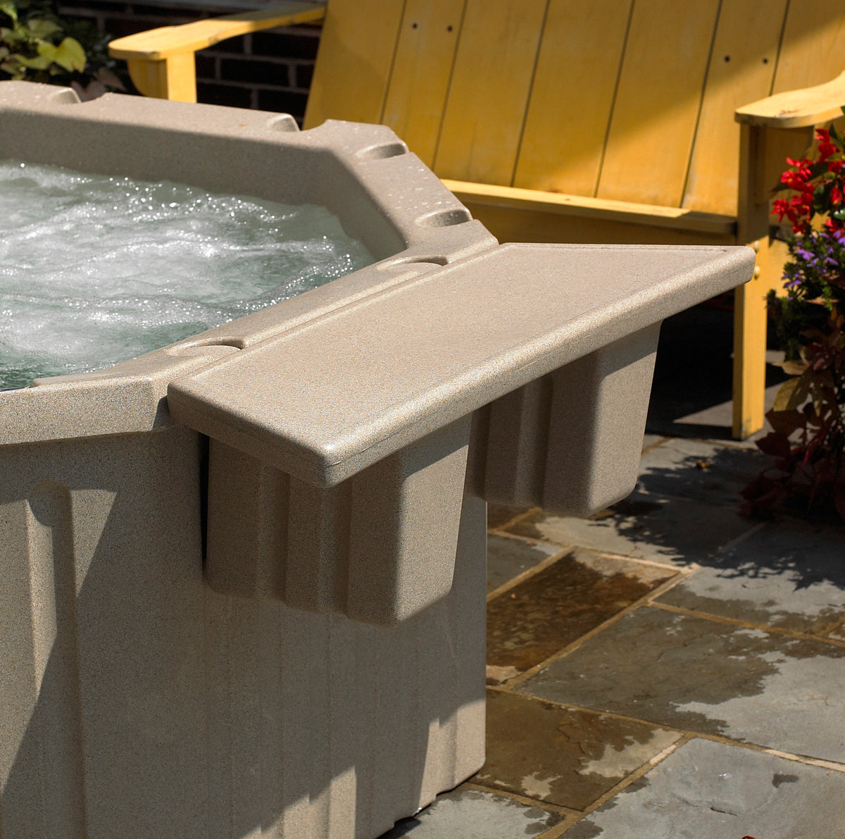 A beige outdoor hot tub with bubbling water and a matching side table extension, shown on a stone patio with a yellow chair in the background.