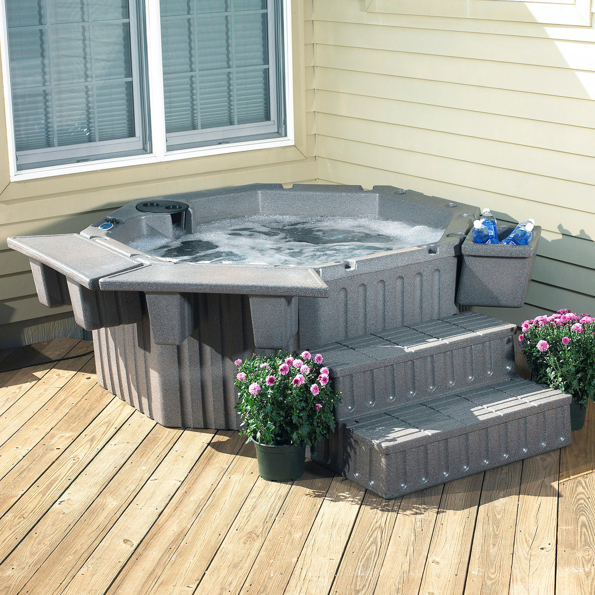 Gray outdoor hot tub with bubbling water, set on a wooden deck beside a house with beige siding. The hot tub includes steps and trays and ice bucket, with potted flowers in the foreground.