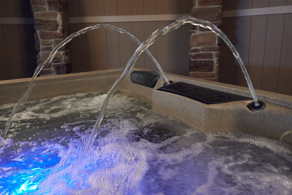 A close-up of a hot tub or spa with bubbling water and two arched jets spraying into the air. The tub has a speckled beige exterior, and blue LED lighting illuminates the water surface.