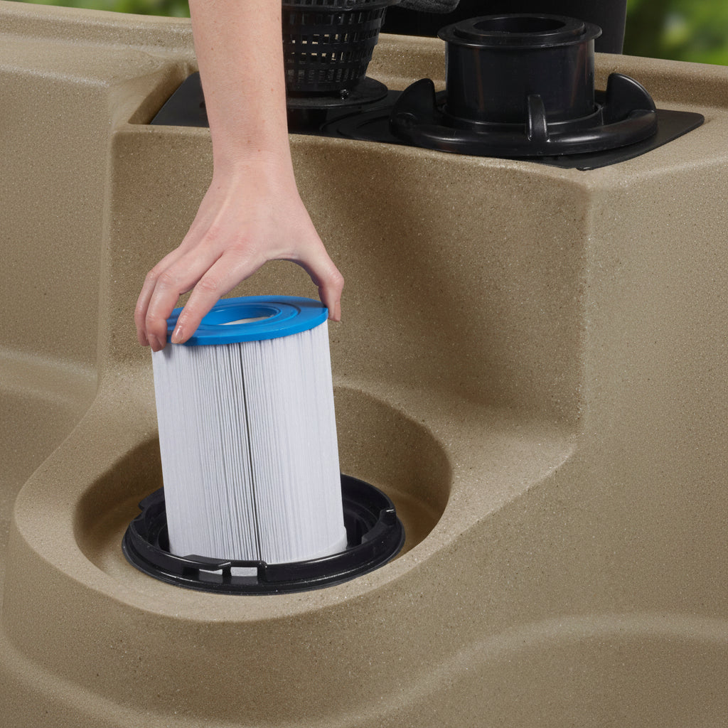 A person's hand is shown removing a white pleated filter cartridge with a blue top from a black circular port in a beige resin spa