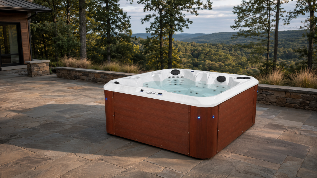A square hot tub with a brown wood-look exterior and white interior, shown on a stone patio with a scenic mountain and forest background.