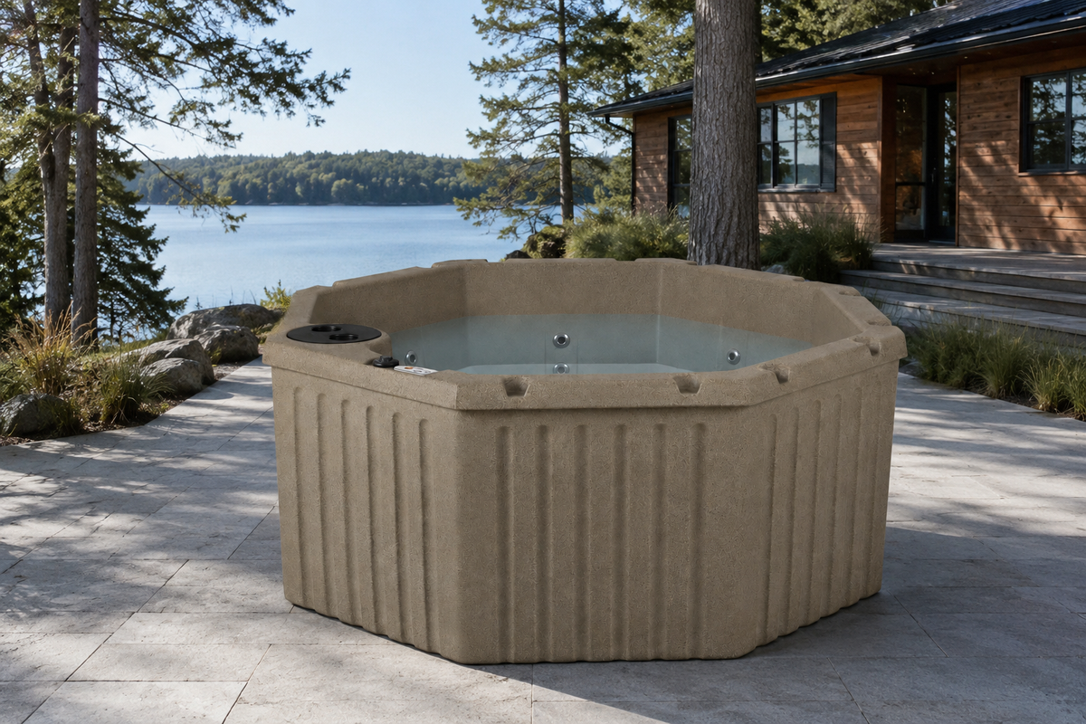 A beige outdoor hot tub with a ribbed exterior and clear water, situated on a stone patio next to a lake with trees and a cabin in the background.