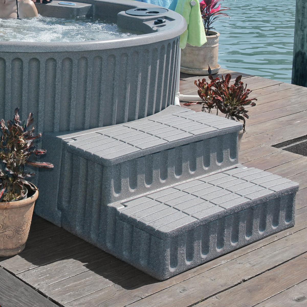 Gray storage steps with adapter for round hot tubs, shown on a deck with hot tub, plants and water