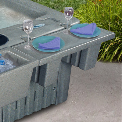 A gray textured hot tub with an integrated tray, shown outdoors on a concrete patio with a person's leg visible in the water and drink accessories placed on the tray.