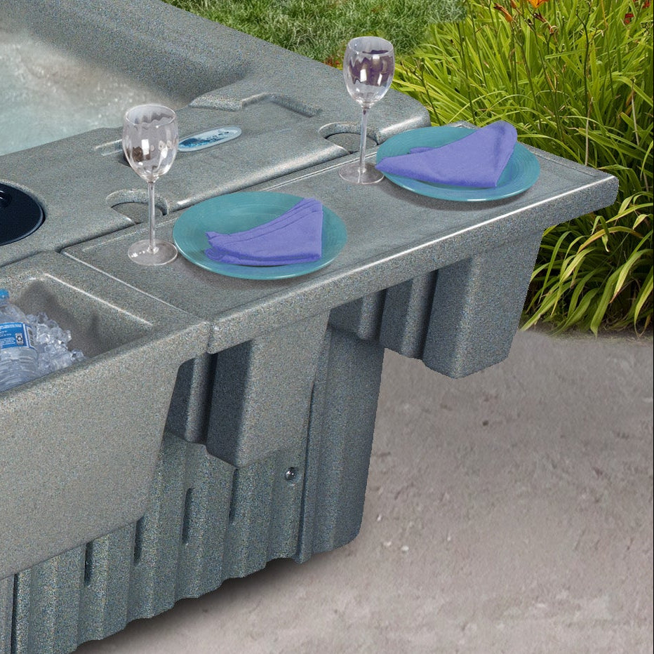 A gray textured hot tub with an integrated tray, shown outdoors on a concrete patio with a person's leg visible in the water and drink accessories placed on the tray.