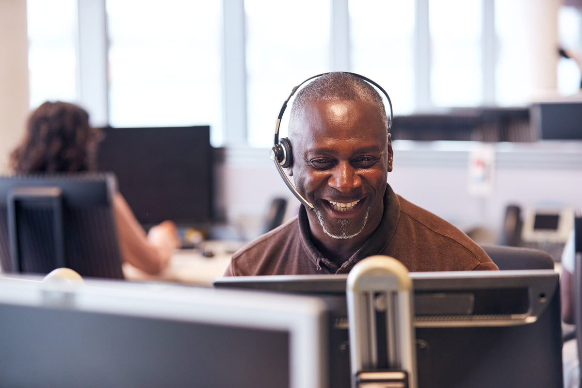 A smiling man wearing a headset sits at a computer desk in a bright office environment.