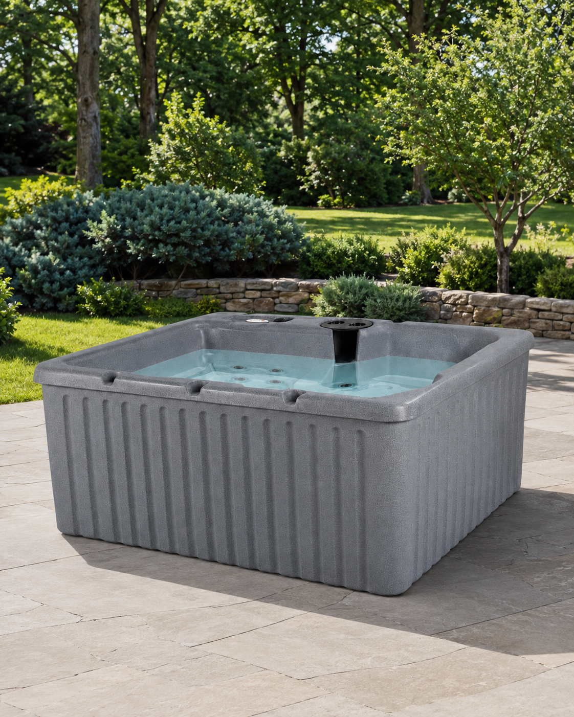 A gray rectangular hot tub with a ribbed exterior and clear water, set on a stone patio in a landscaped garden with trees and shrubs 