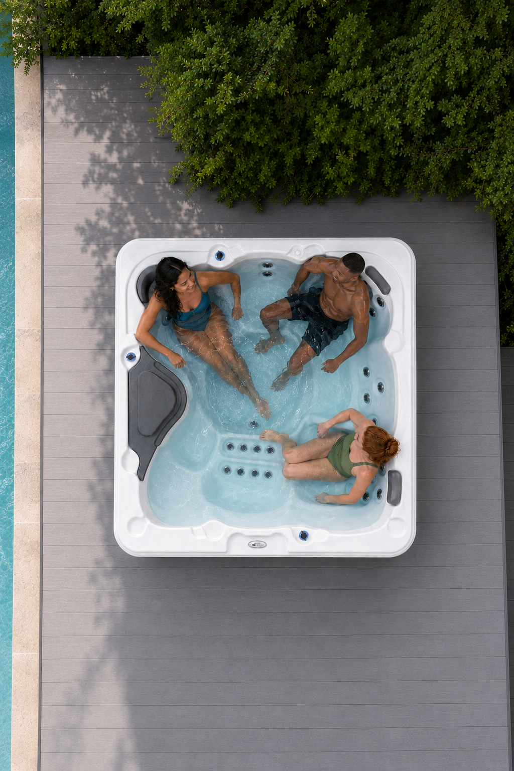 An overhead view of a white outdoor hot tub on a gray deck, with three people relaxing in the water next to a pool and green trees.